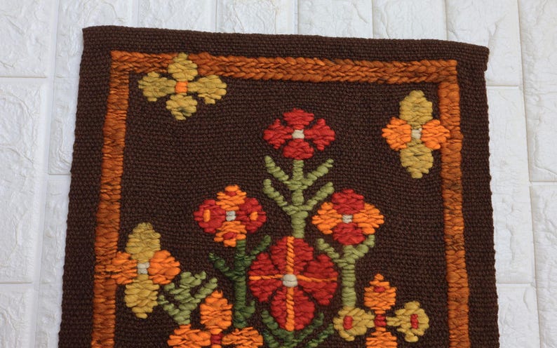 Op de afbeelding: Een bruin vloerkleed met een bloemenpatroon. Het vloerkleed is gemaakt van wol en heeft een franje rand. De bloemen zijn in tinten oranje, rood en geel.