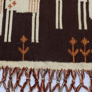 May include: A woven wall hanging with a brown background and cream-colored geometric patterns. The design includes stylized animal figures and floral motifs. The bottom edge features a fringe of brown and cream-colored knotted tassels.
