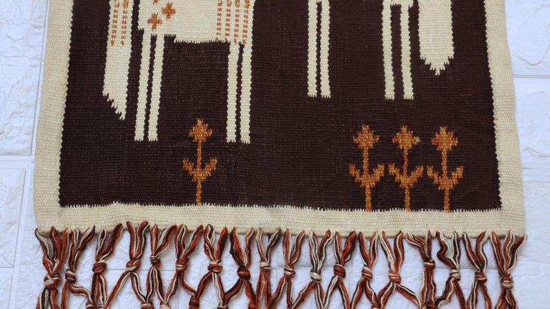 May include: A woven wall hanging with a brown background and cream-colored geometric patterns. The design includes stylized animal figures and floral motifs. The bottom edge features a fringe of brown and cream-colored knotted tassels.
