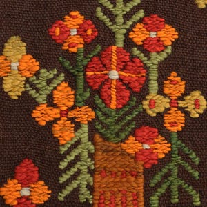 Op de afbeelding: Een bruine geweven wandhanger met een bloemenmotief. De bloemen zijn oranje, rood en geel en zijn gerangschikt in een vaas. De vaas is bruin en heeft een textuur.