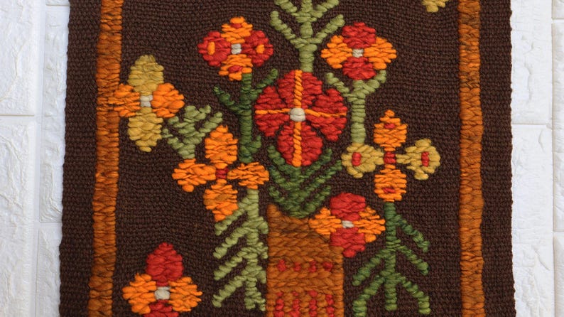Op de afbeelding: Een bruine geweven wandhanger met een bloemenmotief. De bloemen zijn oranje, rood en geel en zijn gerangschikt in een vaas. De vaas is bruin en heeft een textuur.