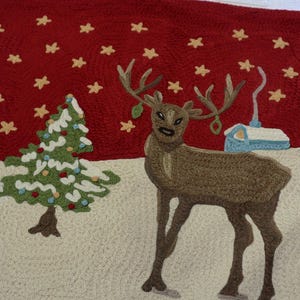 May include: A Christmas-themed rug with a red background and a winter scene. The rug features a reindeer, two Christmas trees, and a small building. The trees are green with white accents, and the reindeer is brown.