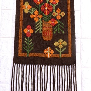 Op de afbeelding: Een bruine geweven wandhanger met een franje rand. De hanger heeft een kleurrijk bloemenmotief met oranje, rode, gele en groene bloemen in een bruine vaas.