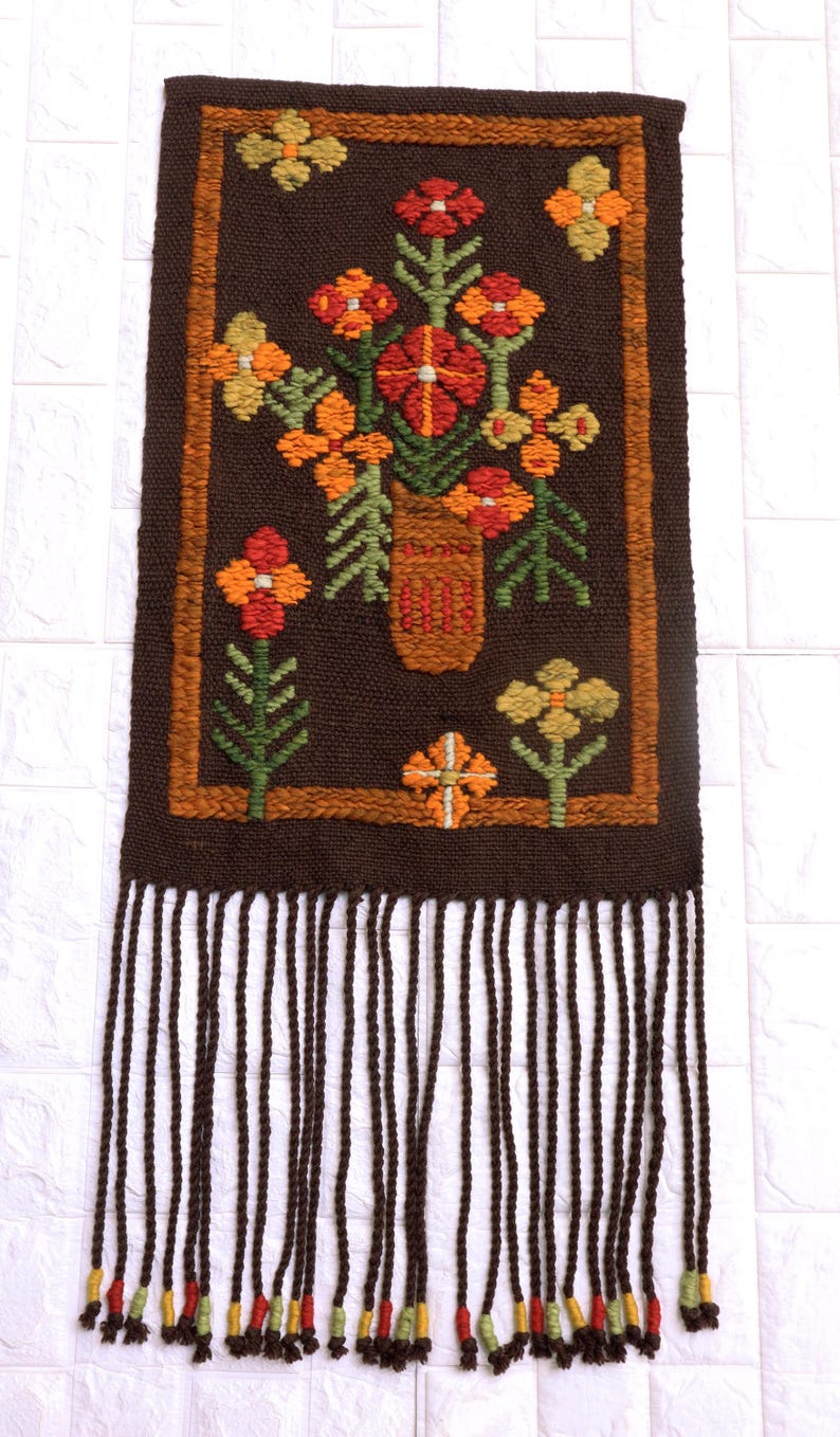Op de afbeelding: Een bruine geweven wandhanger met een franje rand. De hanger heeft een kleurrijk bloemenmotief met oranje, rode, gele en groene bloemen in een bruine vaas.