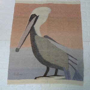 May include: Woven wall hanging featuring a pelican. The artwork depicts the bird in shades of gray, brown, and white against a backdrop of orange, blue, and beige. The piece is signed in the lower left corner.