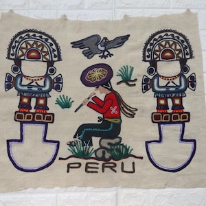 Op de afbeelding: Een beige textiele wandhanger met geborduurde figuren en het woord "PERU". Het ontwerp toont twee gestileerde figuren, een zittende persoon die fluit speelt en een vogel. De figuren zijn in rood, blauw en bruin.