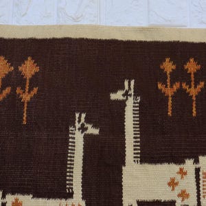 May include: A woven wall hanging featuring a brown background with two giraffes and stylized floral elements in tan and orange. The border is a lighter tan color.