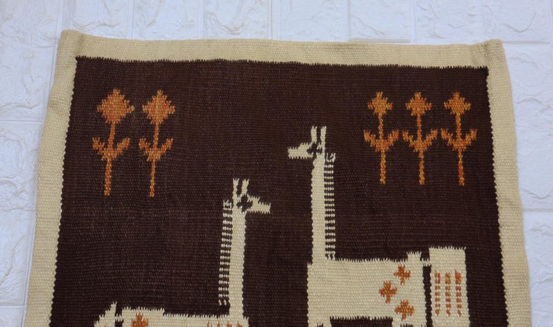 May include: A woven wall hanging featuring a brown background with two giraffes and stylized floral elements in tan and orange. The border is a lighter tan color.