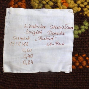 Op de afbeelding: Een wit label met zwarte tekst die "Wierzbicka Stanislawa Swigtek Danuta Sumak "Bukiet" 13/12/142 64-Buk 0,60 0,40 0,24" luidt. Het label is bevestigd aan een bruin geweven tapijt met oranje en gele bloemenmotieven.