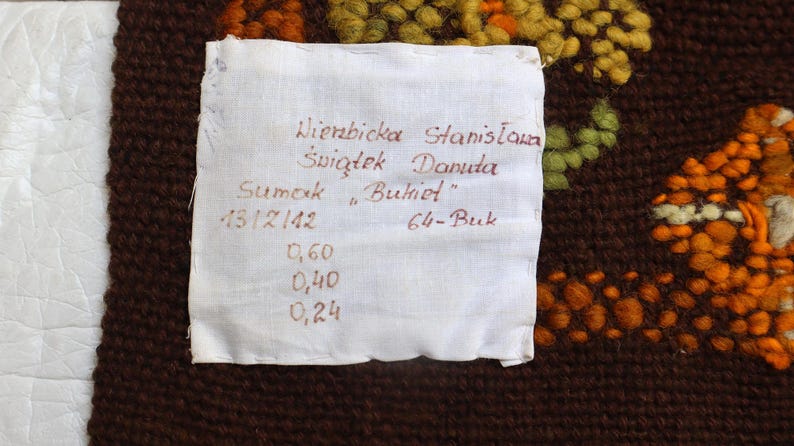 Op de afbeelding: Een wit label met zwarte tekst die "Wierzbicka Stanislawa Swigtek Danuta Sumak "Bukiet" 13/12/142 64-Buk 0,60 0,40 0,24" luidt. Het label is bevestigd aan een bruin geweven tapijt met oranje en gele bloemenmotieven.