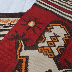 Puede incluir: Textil tejido a mano con fondo rojo y patrones geométricos en blanco, marrón y naranja. El diseño incluye formas solares y figuras estilizadas. El segundo textil tiene rayas marrones y blancas.