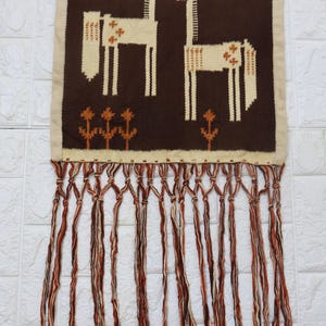 May include: A woven wall hanging featuring two stylized giraffes in cream and brown. The giraffes are set against a dark brown background with cream-colored floral accents. The piece has a cream border and a fringe of brown, cream, and orange strands.