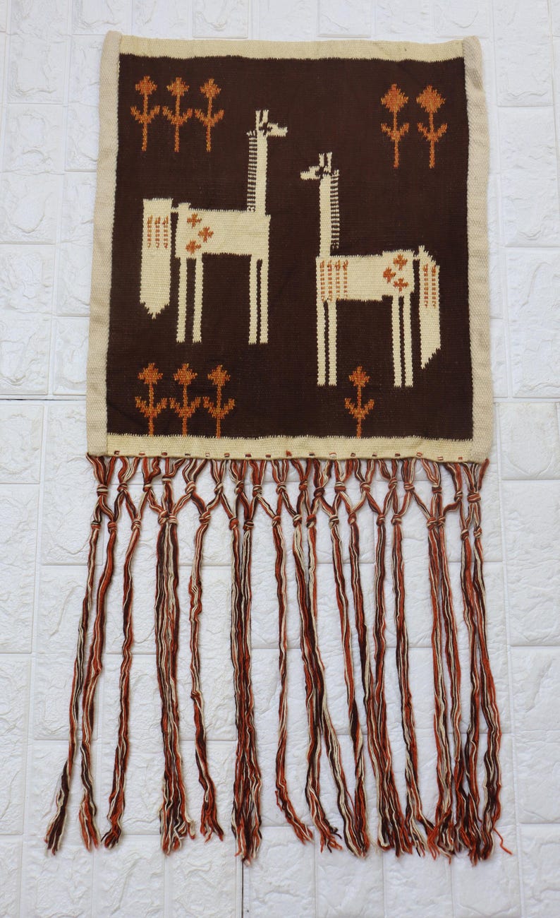 May include: A woven wall hanging featuring two stylized giraffes in cream and brown. The giraffes are set against a dark brown background with cream-colored floral accents. The piece has a cream border and a fringe of brown, cream, and orange strands.