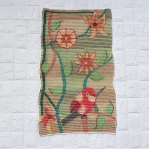May include: A rectangular woven wall hanging featuring a tan background with a repeating horizontal ribbed texture. The design displays a red bird perched on a branch, surrounded by green leaves and orange and red floral motifs with dark outlines.