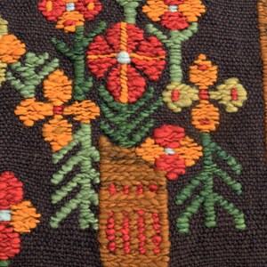 Op de afbeelding: Een close-up van een bruine geweven wandhanger met een kleurrijk bloemenontwerp. De bloemen zijn rood, oranje en geel, en de bladeren zijn groen. De bloemen zijn gerangschikt in een vaas, die ook geweven en bruin is.