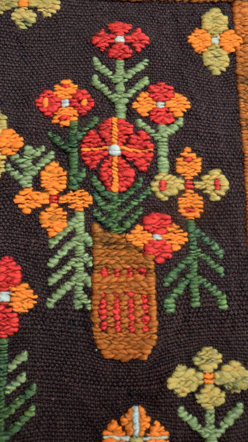 Op de afbeelding: Een close-up van een bruine geweven wandhanger met een kleurrijk bloemenontwerp. De bloemen zijn rood, oranje en geel, en de bladeren zijn groen. De bloemen zijn gerangschikt in een vaas, die ook geweven en bruin is.