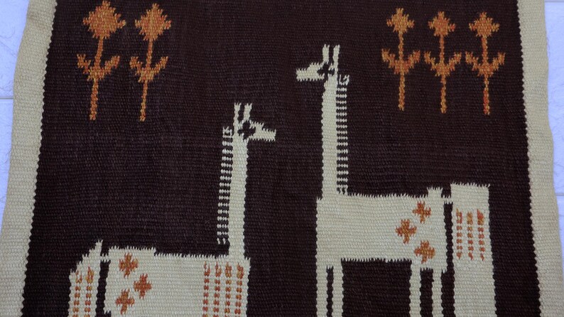 May include: Woven textile featuring two giraffes and stylized trees in brown, cream, and orange. The giraffes have long necks and are set against a dark brown background. The border is cream-colored.