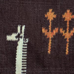 May include: Close-up of a woven textile with a dark brown background. The design features white giraffe silhouettes and orange floral-like patterns. The textile has a textured appearance and a white border.