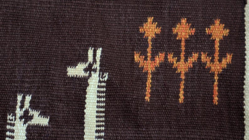 May include: Close-up of a woven textile with a dark brown background. The design features white giraffe silhouettes and orange floral-like patterns. The textile has a textured appearance and a white border.