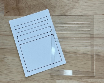 May include: A clear plastic stencil with lines and a rectangle for creating a simple card or journal page. The stencil is placed on top of a white card with lines and a rectangle drawn on it.