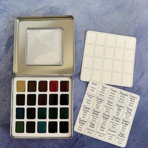 May include: A watercolor paint set in a silver tin with a clear window. The set includes 16 vibrant colors, with color names on a separate card. Additional blank cards are included for mixing. The set is ideal for artists.