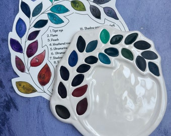 Può includere: Una tavolozza da artista in ceramica bianca con una tabella dei colori a forma di foglia. La tavolozza ha vari pozzetti di vernice riempiti di colori scuri e vibranti. La tabella dei colori mostra campioni di colore numerati con nomi come "Tiger eye" e "Flame".