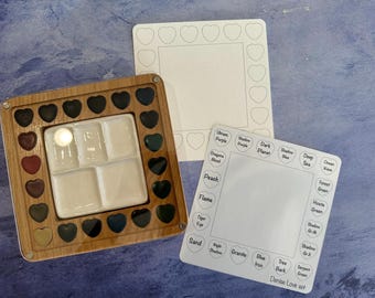 Könnte beinhalten: Eine hölzerne Aquarellpalette mit einem weißen Keramik-Mischbereich und herzförmigen Farbnäpfchen. Zwei weiße Karten mit Herzrändern und Farbbezeichnungen wie "Pfirsich" und "Granit" sind ebenfalls vorhanden. Der Text "Denise Love set" ist sichtbar.