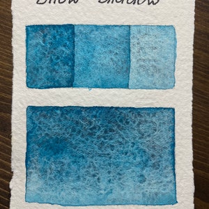 May include: A watercolor painting of a blue color gradient with the words "Snow Shadow" written at the top.