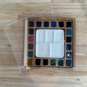 May include: A wooden watercolour paint palette with a clear acrylic lid. The palette has a white ceramic mixing area and contains various colours of paint. The palette is square and sits on a wooden surface.