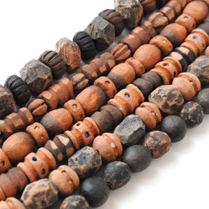 Op de afbeelding: Close-up van verschillende strengen houten kralen in diverse vormen en maten. De kralen zijn voornamelijk bruin en zwart, met enkele blauwe accenten. De kralen hebben een rustieke, handgemaakte uitstraling, wat duidt op een natuurlijke of bohemien stijl.