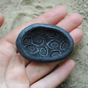 May include: A dark grey, oval ceramic dish featuring an eye-like pattern carved into its interior. The dish is held in a hand, highlighting the detailed texture and design. The background is a sandy surface.