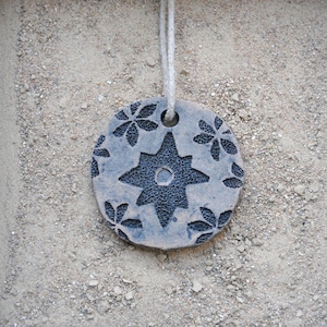 Può includere: Un ciondolo rotondo in ceramica grigia con un motivo a stella e floreale. Il ciondolo è sospeso da un cordino bianco. La stella al centro ha un aspetto strutturato, e i fiori sono distanziati.