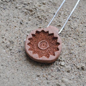 Può includere: Una collana con pendente color terracotta con un design circolare dettagliato. Il pendente è sospeso a una delicata catena d'argento. L'intricato motivo sul pendente ricorda un fiore o un sole stilizzato, su uno sfondo sabbioso.