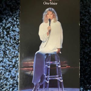 May include: A VHS tape case featuring a black and white photo of Barbra Streisand singing into a microphone. The text on the case reads "Barbra Streisand One Voice".