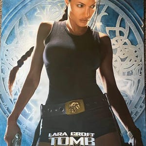 May include: A movie poster for "Lara Croft: Tomb Raider" featuring a woman in a black tank top and shorts, holding a gun. The poster has a blue and silver background with the movie title prominently displayed.