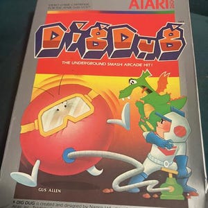 May include: A vintage Atari 2600 video game cartridge for Dig Dug. The box features the game's logo in orange and blue, with a red background. The cover art shows a character with a pump and a monster.
