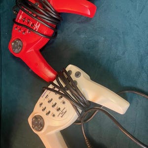 May include: Two retro red and off-white game controllers with black cords and connectors. The red controller has several buttons and a directional pad. The off-white controller has the text "Explore Super Driving for Saturn".