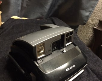 May include: A silver and black Polaroid camera with a strap. The camera has a built-in flash and a viewfinder.