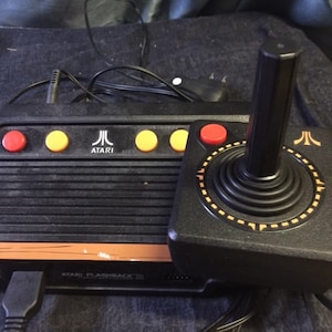 May include: A black Atari Flashback 8 console with a joystick. The console has a black and orange design with the Atari logo and the text "ATARI FLASHBACK 8" printed on the front. The joystick has a black base with a black stick and a yellow ring around the base.
