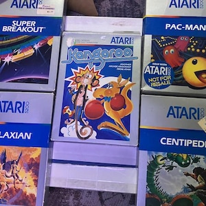 Peut inclure: Plusieurs cartouches de jeux vidéo Atari 5200. Les titres incluent "Super Breakout", "Kangaroo", "Pac-Man", "Galaxian" et "Centipede". Les boîtes présentent des illustrations colorées et le logo Atari.