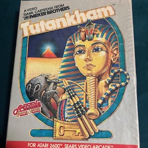 May include: A vintage video game cartridge for the Atari 2600, featuring the game "Tutankham." The box art showcases a stylized depiction of Tutankhamun with a golden headdress, snakes, and hieroglyphic elements. The "Arcade Game Series" logo is visible.