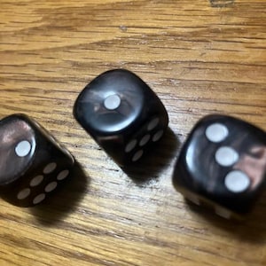 May include: Three black dice with white dots are arranged on a wooden surface. The dice are slightly translucent, with a marbled effect. The dots are clearly visible, indicating the numbers on each die. The dice are likely used for games.