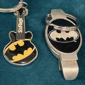 May include: Two keychains: one shaped like a guitar with a black and yellow Batman logo and the text "Six Flags", and a silver bottle opener with a black and white Batman logo.
