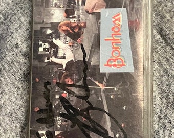 May include: A cassette tape with a black and white photo of a band on the cover. The band is standing in a room with a checkered floor. The tape is signed by the band, "Bon Jovi".