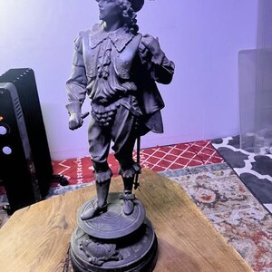 May include: A detailed bronze statue of a man in historical attire, including a hat with a feather, a ruffled collar, and a vest. The statue is mounted on a round, tiered base with decorative elements. The statue is on a wooden surface.