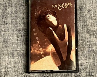May include: A cassette tape case with a brown background and a photo of Mariah Carey. The text "Mariah Carey" is in gold lettering at the top of the case. The text "Emotions" is in gold lettering on the left side of the case. The photo shows Mariah Carey in a white tank top with her hair blowing in the wind.
