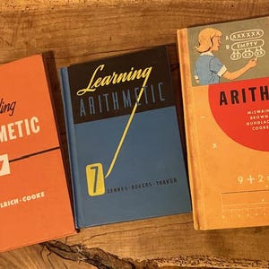 May include: Three vintage arithmetic textbooks. The books are orange and blue, with titles like "Understanding Arithmetic" and "Learning Arithmetic." One book features a diagram of a woman teaching math. The books are numbered 3 and 7.