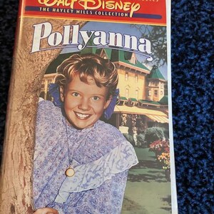 May include: A VHS tape featuring the Walt Disney Film Classics: The Hayley Mills Collection, with the title "Pollyanna" in large, colorful letters. The cover features a young girl with blonde hair, wearing a blue and white floral dress, smiling and holding a tree trunk. A large house is visible in the background.