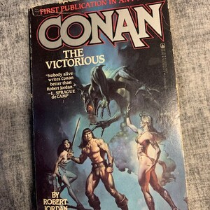 May include: A paperback book cover featuring a fantasy illustration of Conan the Barbarian, a muscular man with a sword, standing in front of a large, shadowy creature. The book title is "Conan the Victorious" and the author is Robert Jordan. The cover also includes a quote from L. Sprague de Camp.