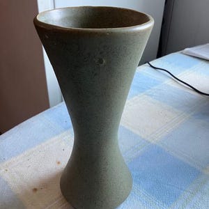 May include: A speckled, olive-green ceramic vase with a flared top and a rounded base. The vase has a smooth, matte finish and a classic hourglass shape. The vase is approximately 8 inches tall.